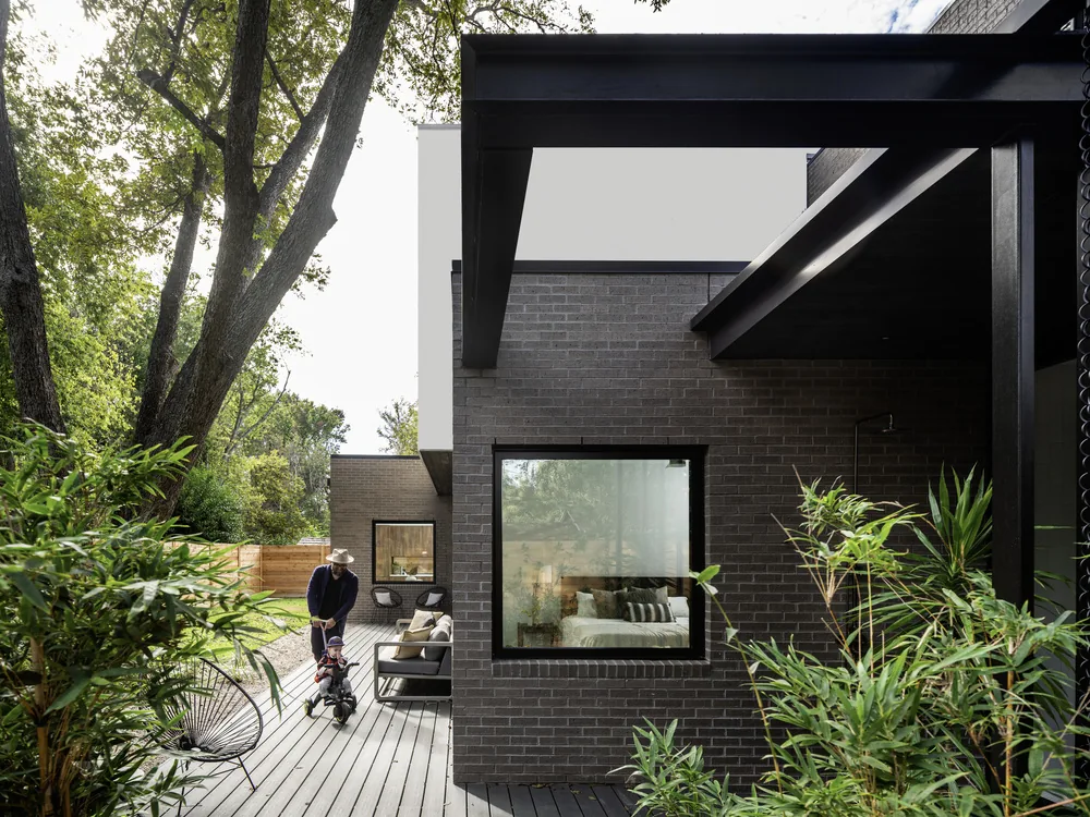 Modern brick home with black window frames, covered patio, and wooden deck visible, featuring outdoor seating and greenery.