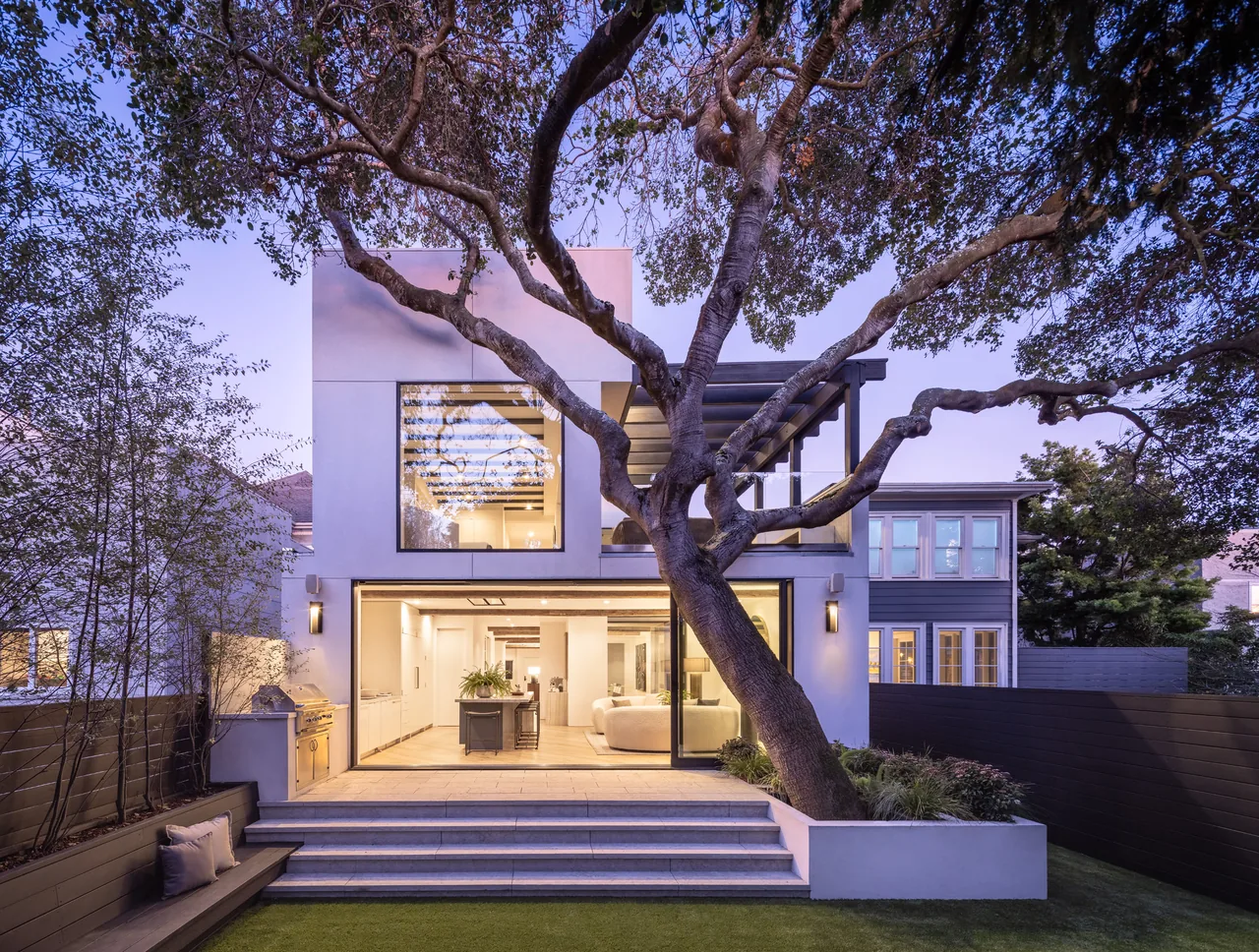 Modern two-story house exterior with large windows and an outdoor seating area beneath a mature tree at dusk.