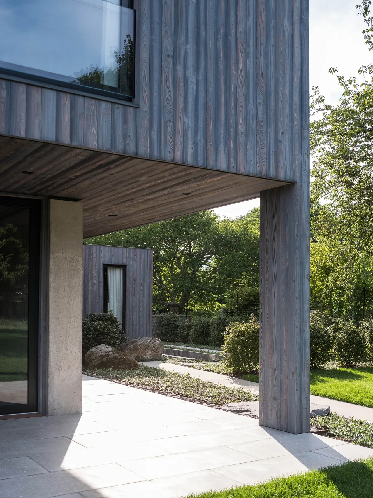 The house features gray vertical wood siding and an overhanging section supported by a wood-clad column, with a paved patio area.