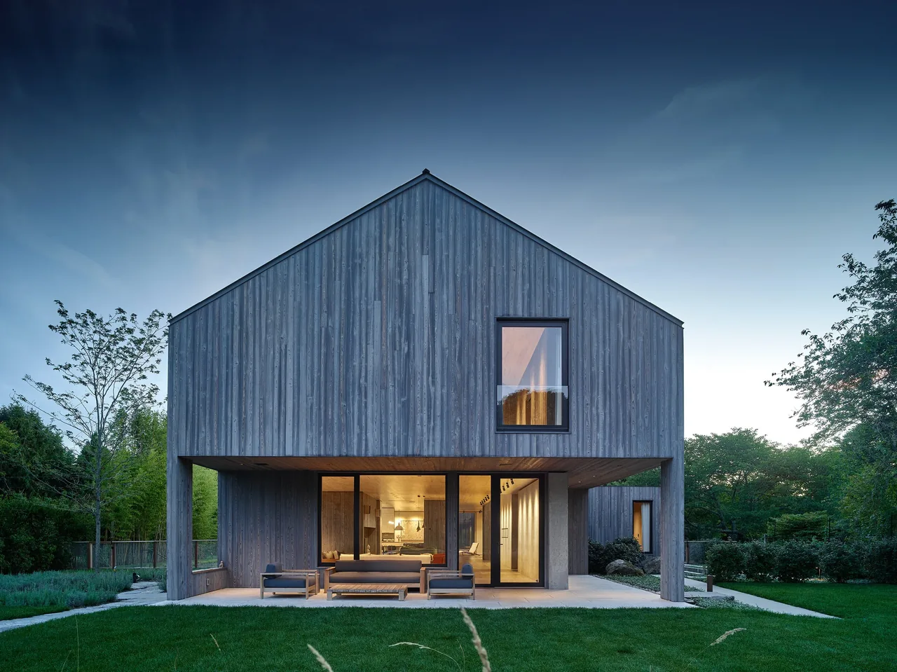 Rear view of the modern wood-clad house showing covered patio and glass sliding doors opening to the living area.