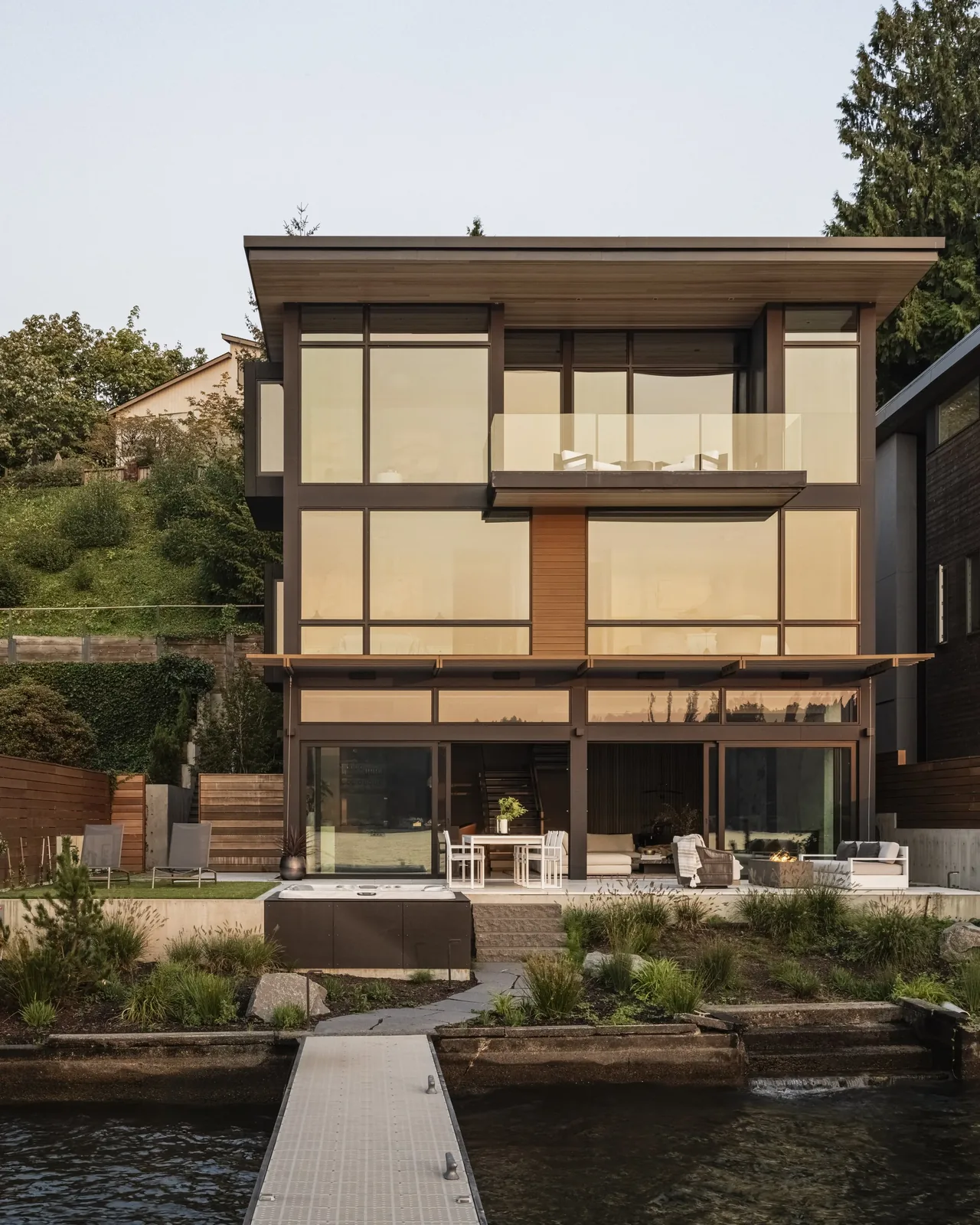 Rear exterior view of a modern three-story house with large glass windows and a dock extending to the water.