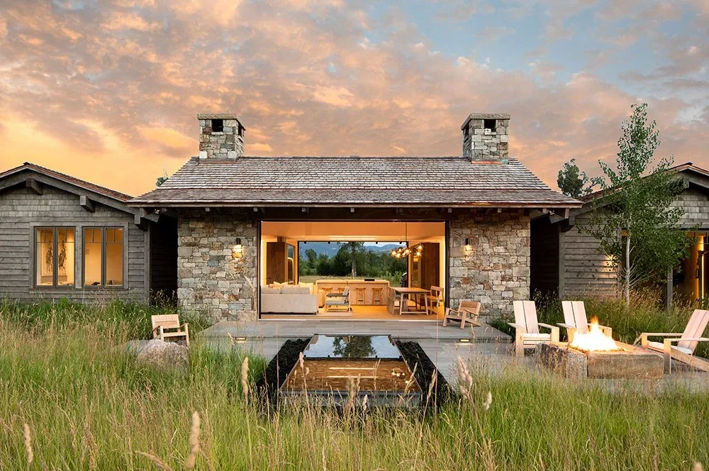 Stone and wood house with open living room and fireplace surrounded by tall grass at sunset.