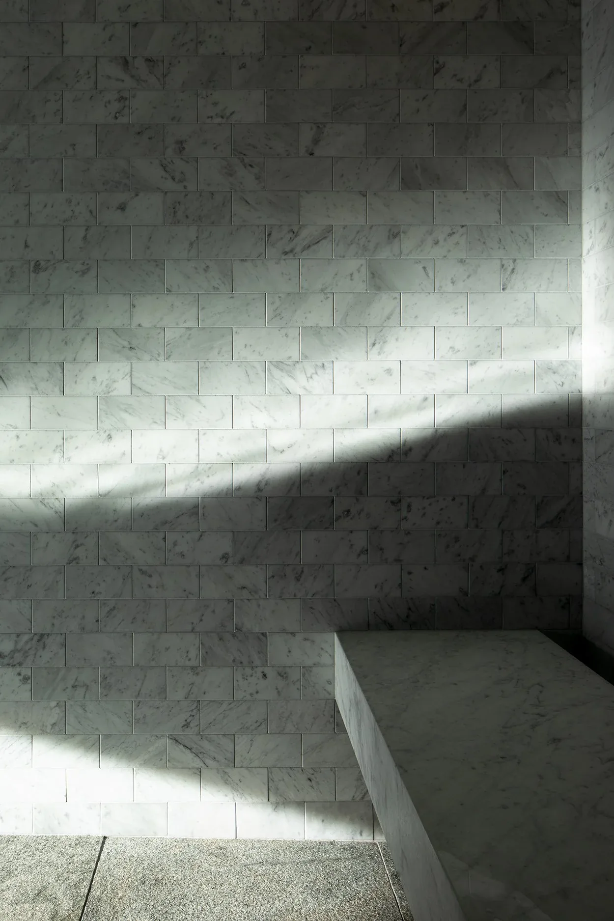 Marble tiles cover the wall and bench of a minimalist interior space with light and shadow play.
