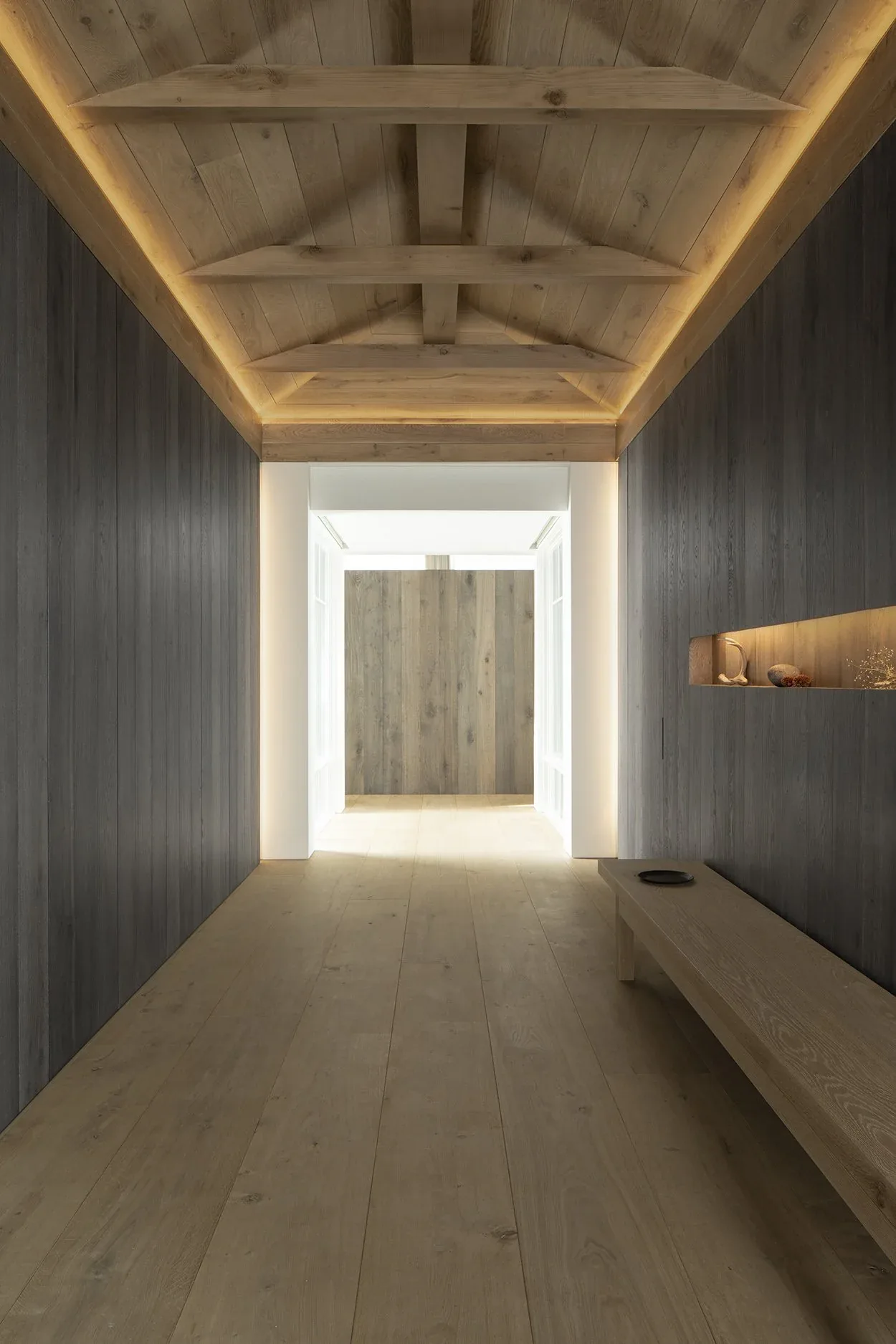 Entry hallway with vaulted wood ceiling, dark wood walls, light wood plank floor, and a lighted niche above a bench.