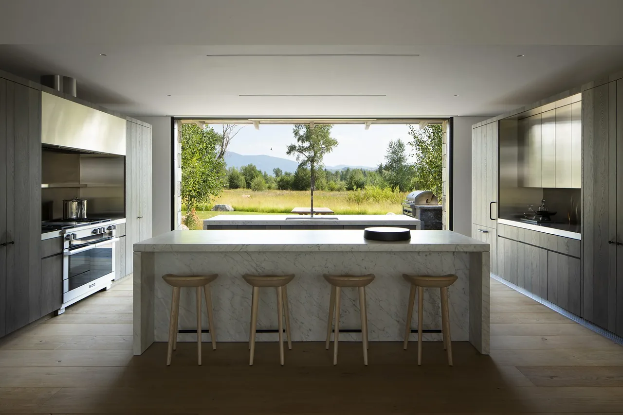 Kitchen with marble island and wood cabinetry opening to outdoor dining and landscape at Junegrass House.