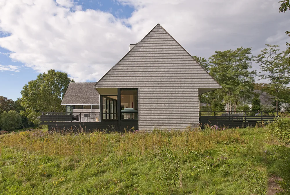 Side view of a residential house with wood shingle siding and a gable roof, surrounded by grassy landscape.