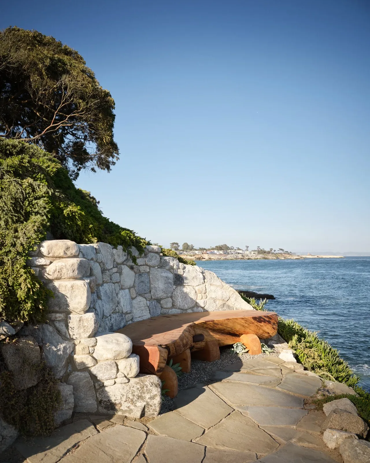 A natural wood bench with a stone wall backdrop sits on a stone-paved patio facing the ocean at Pacific Promontory.
