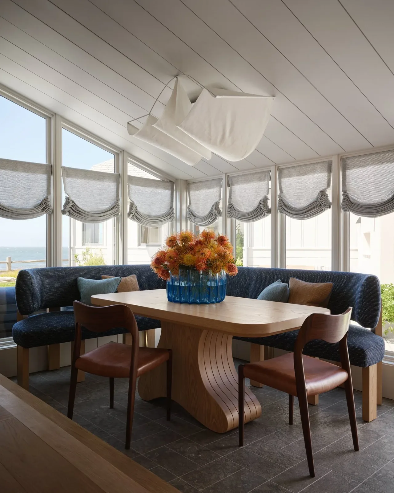 Dining nook with a custom wooden table, curved blue banquette seating, and soft gray Roman shades covers windows with ocean views.