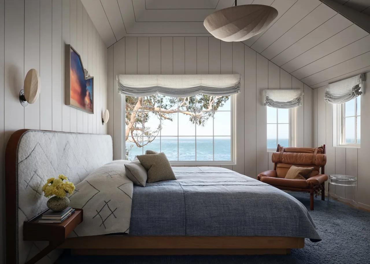 Bedroom features vaulted ceiling, large picture window overlooking the ocean, and leather lounge chair by smaller windows with roman shades.