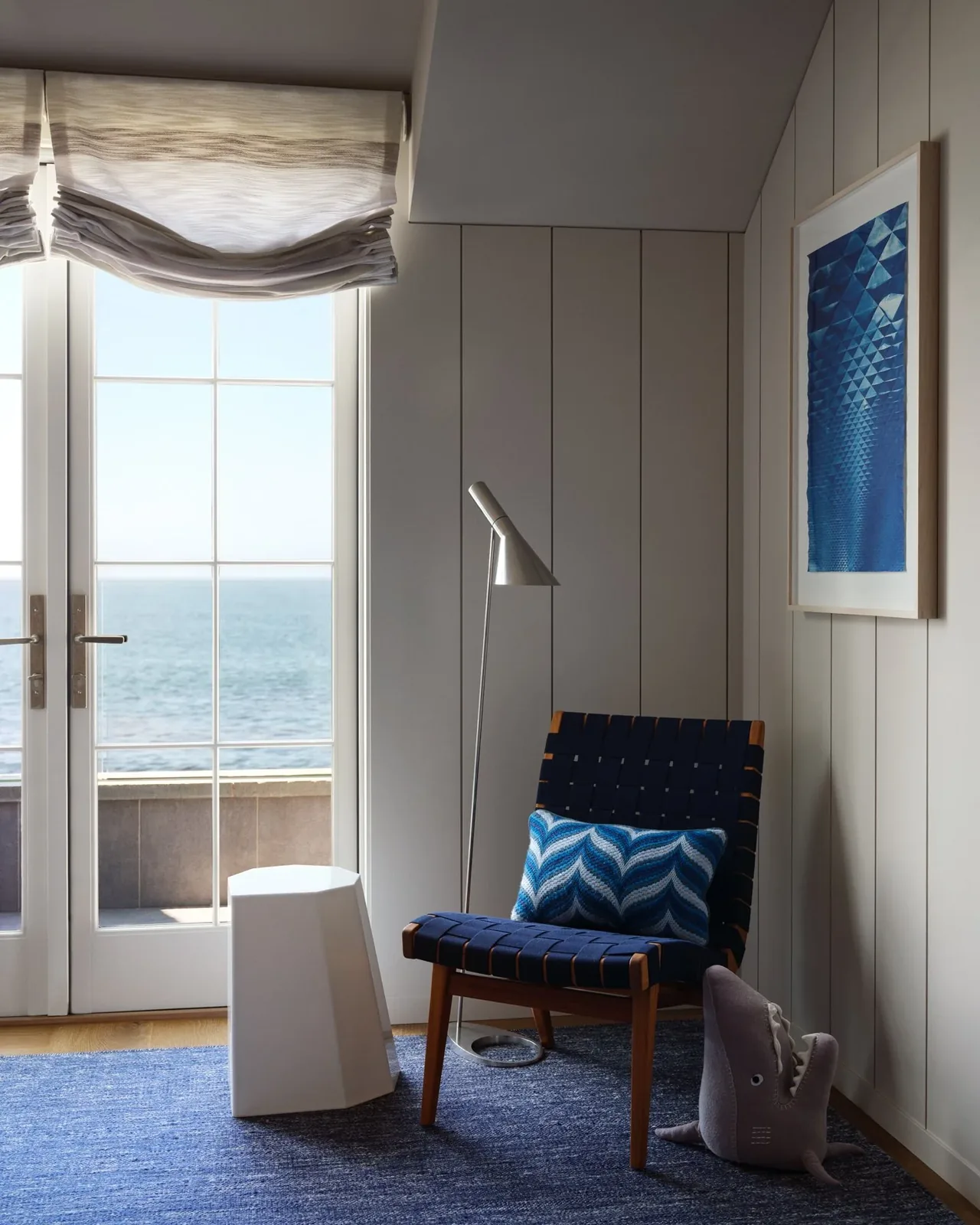 Coastal living space corner features a woven blue chair, blue patterned pillow, floor lamp, and ocean view through French doors.