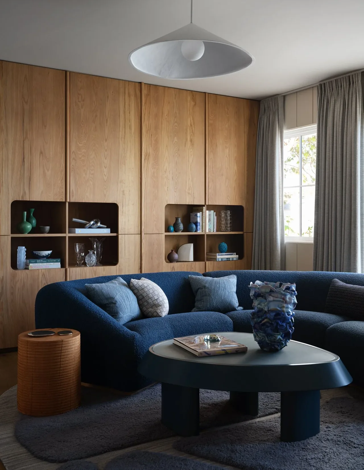Living room features a curved blue sofa, built-in wood shelving with decor, and a central dark coffee table under a conical pendant light.
