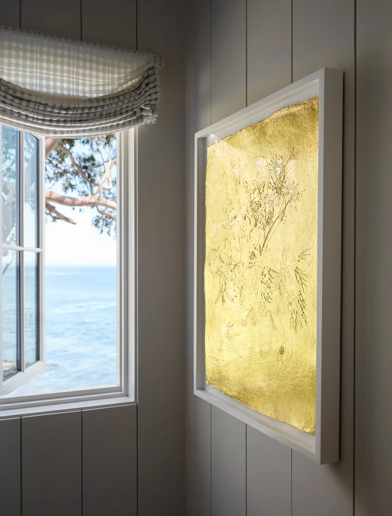Room corner features a checked Roman shade over an open window with ocean view and a backlit golden floral artwork.