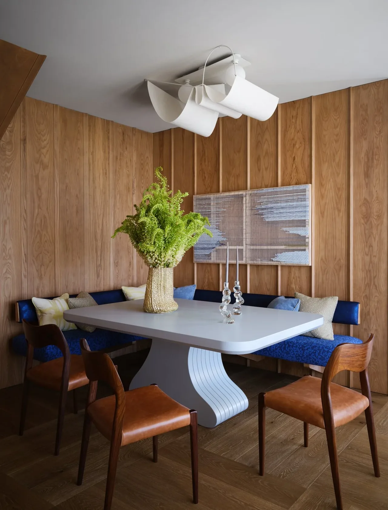 A curved blue upholstered banquette surrounds a white pedestal dining table in a wood-paneled nook with a modern ceiling light and abstract wall art.