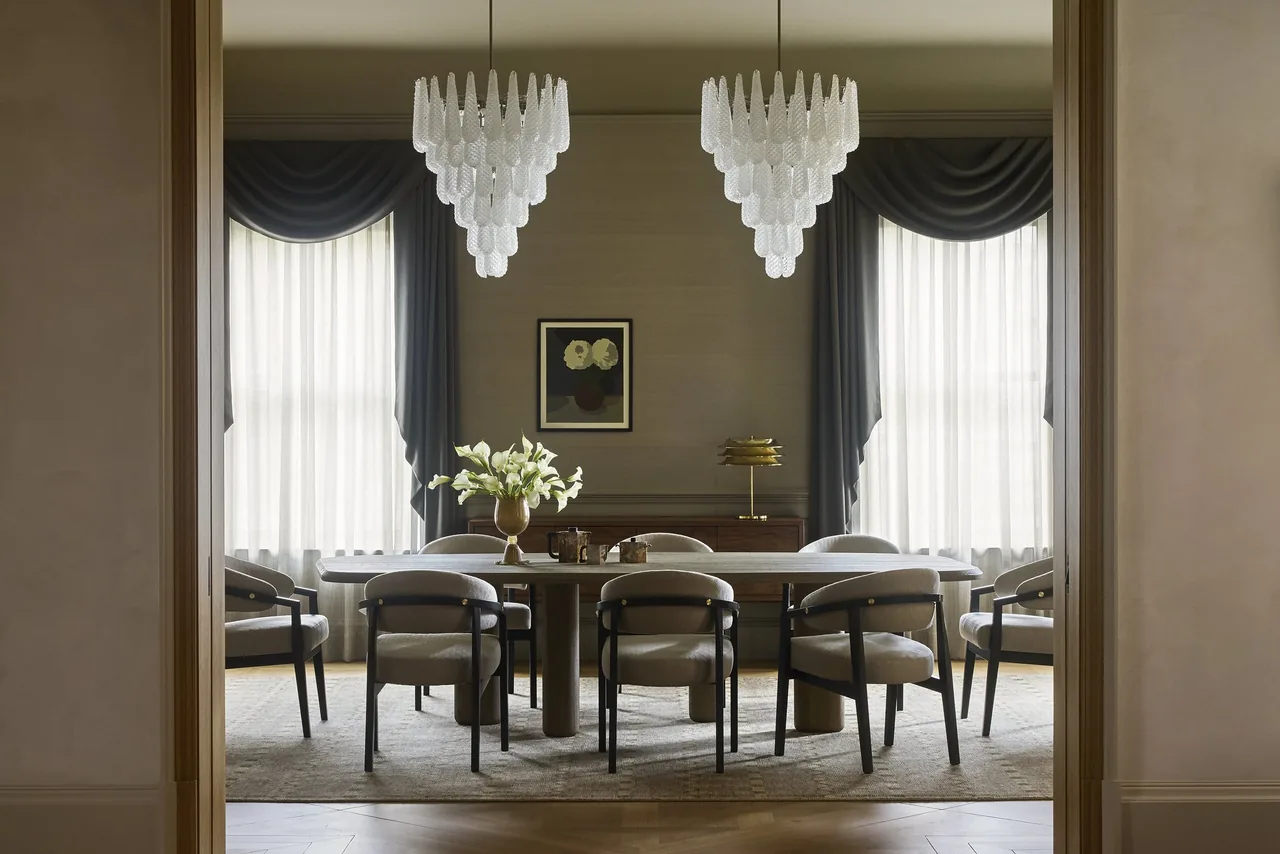 Dining room featuring a large table with upholstered chairs, two tiered chandeliers, and symmetrical window treatments.