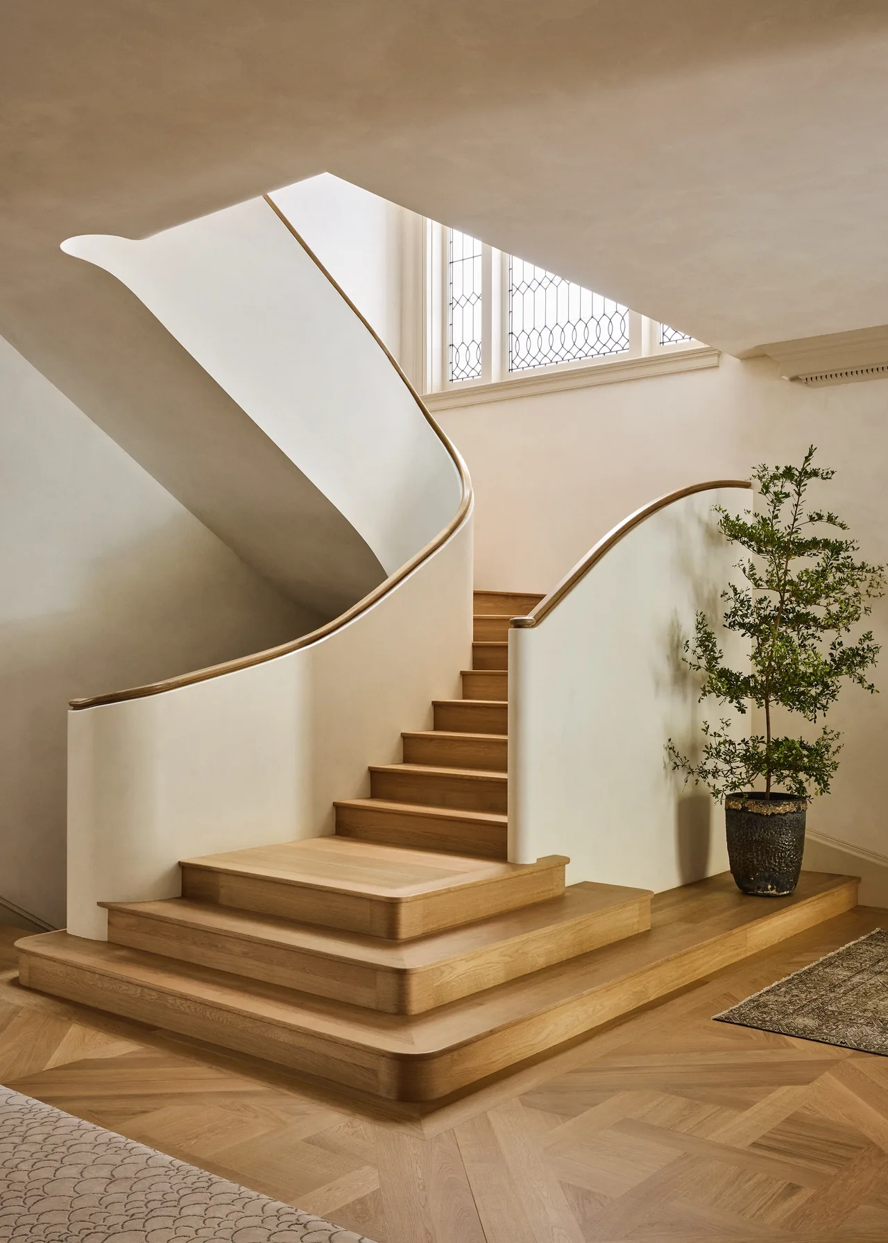A gently curving wooden staircase with a white smooth balustrade and leaded glass windows above illuminates the stairwell.