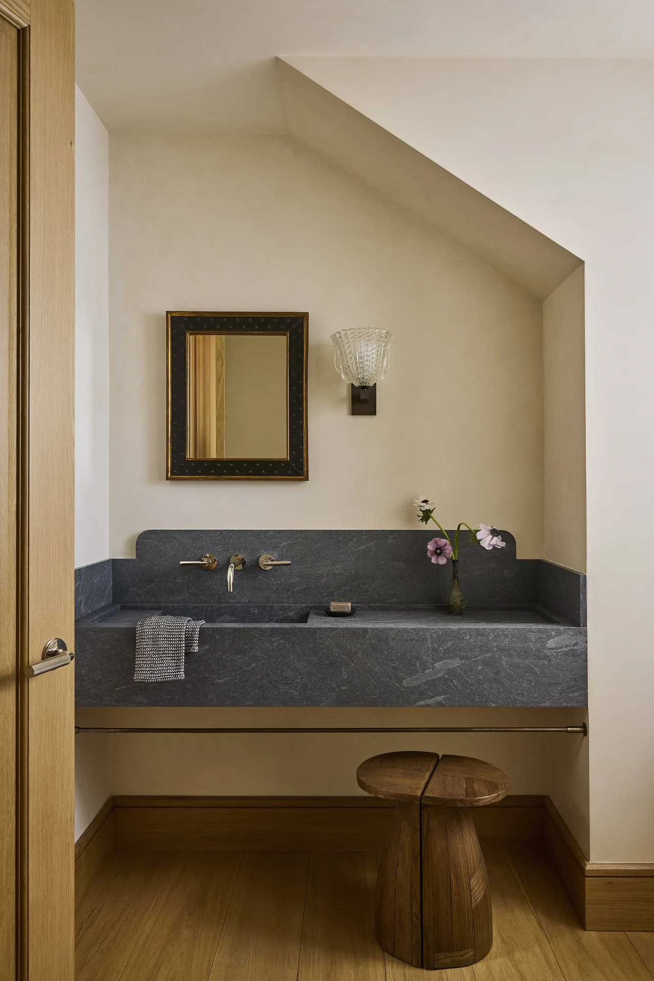 Wall-mounted faucets above a dark gray stone bathroom sink fill a minimalist space with wood floors and a small wooden stool.