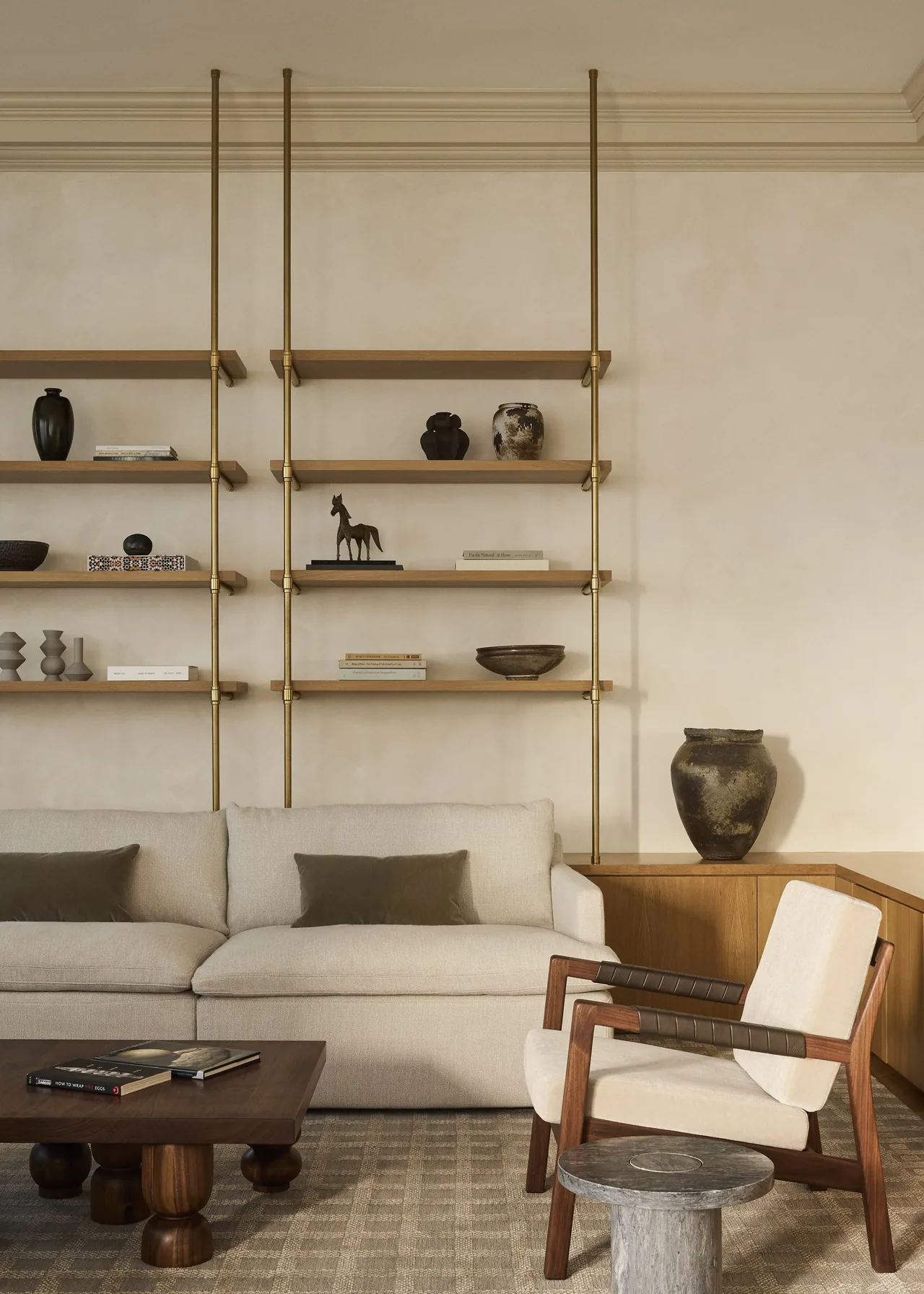 A modern living room features a beige sofa, wooden armchair, and open wood shelving with brass supports.