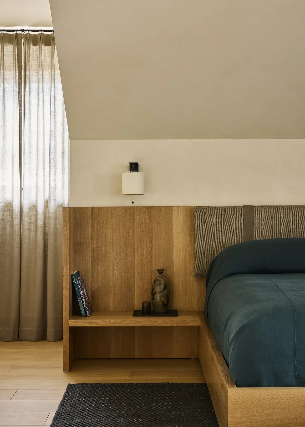 A wooden nightstand is integrated with a bed frame featuring a beige cushioned headboard and a wall-mounted light fixture.