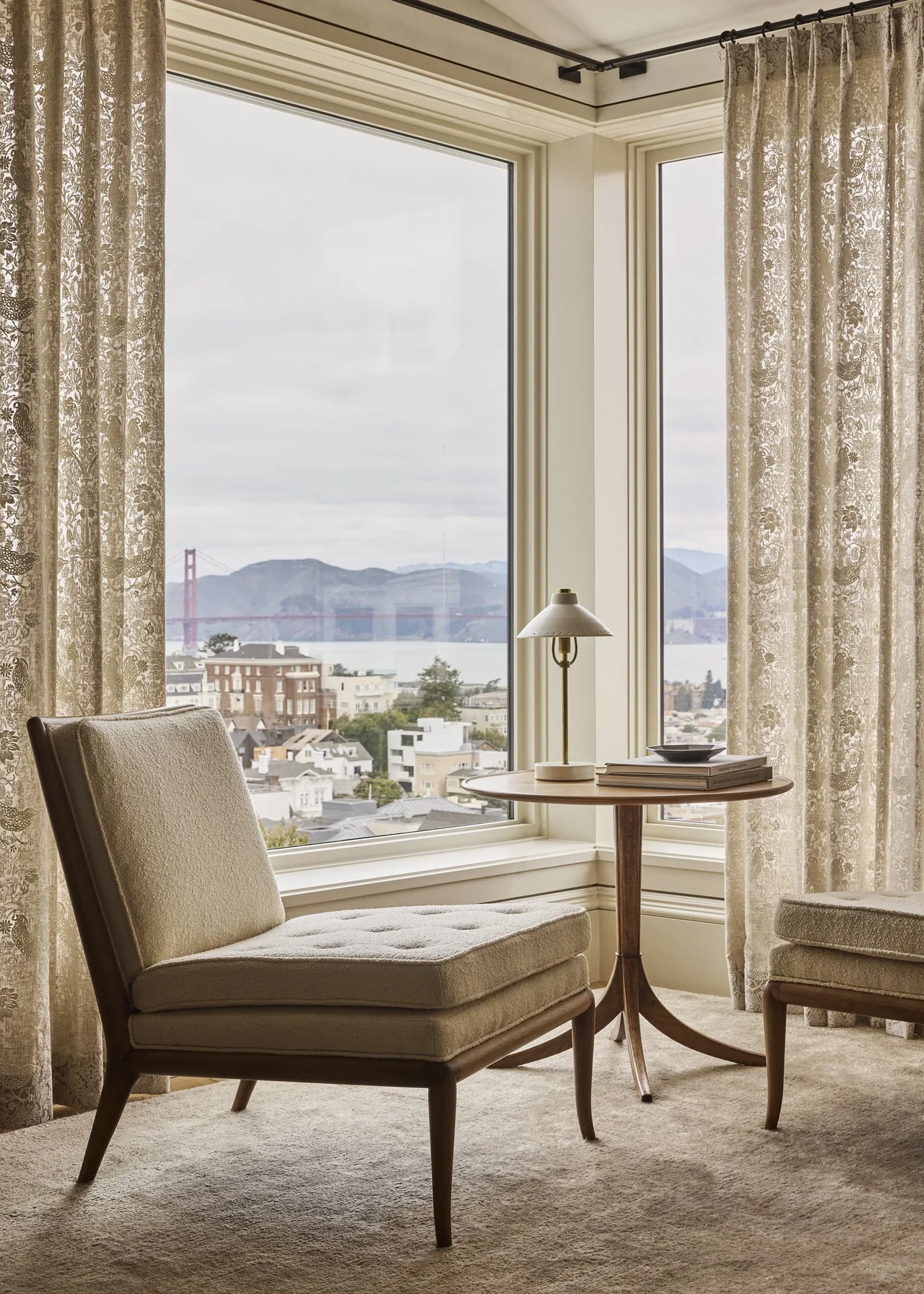 Corner sitting area with beige cushioned chair and table beside a large window overlooking the city and bay with the Golden Gate Bridge visible.