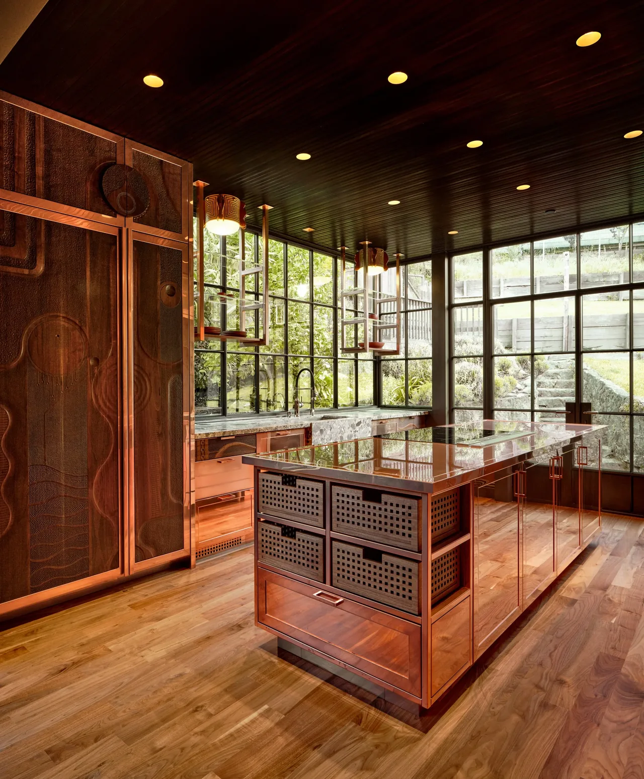 The Gallery House kitchen features reflective copper cabinetry and a central island with expansive grid-style windows offering garden views.