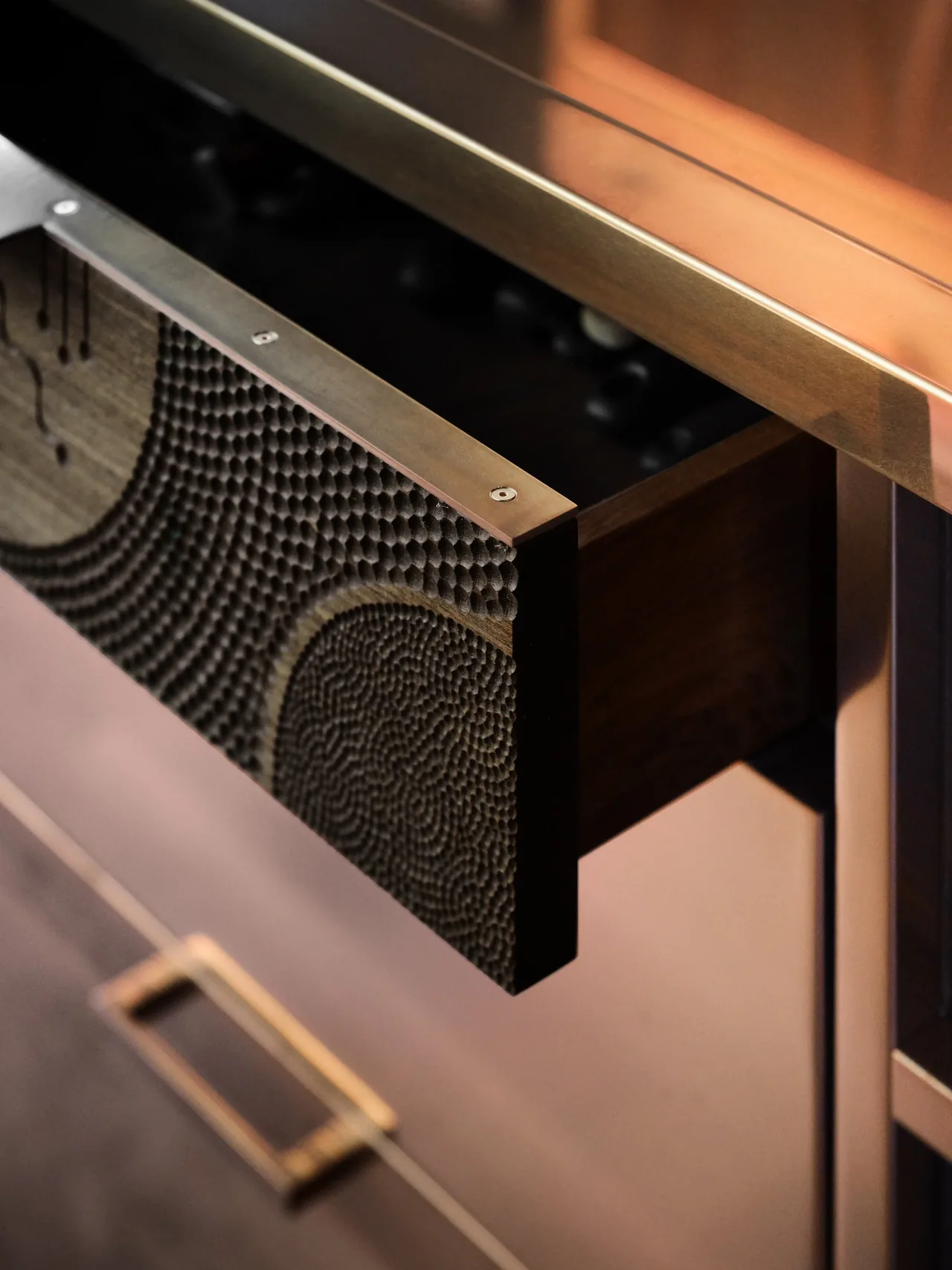 Open drawer showing textured circular wood panel on drawer front and copper kitchen cabinetry.
