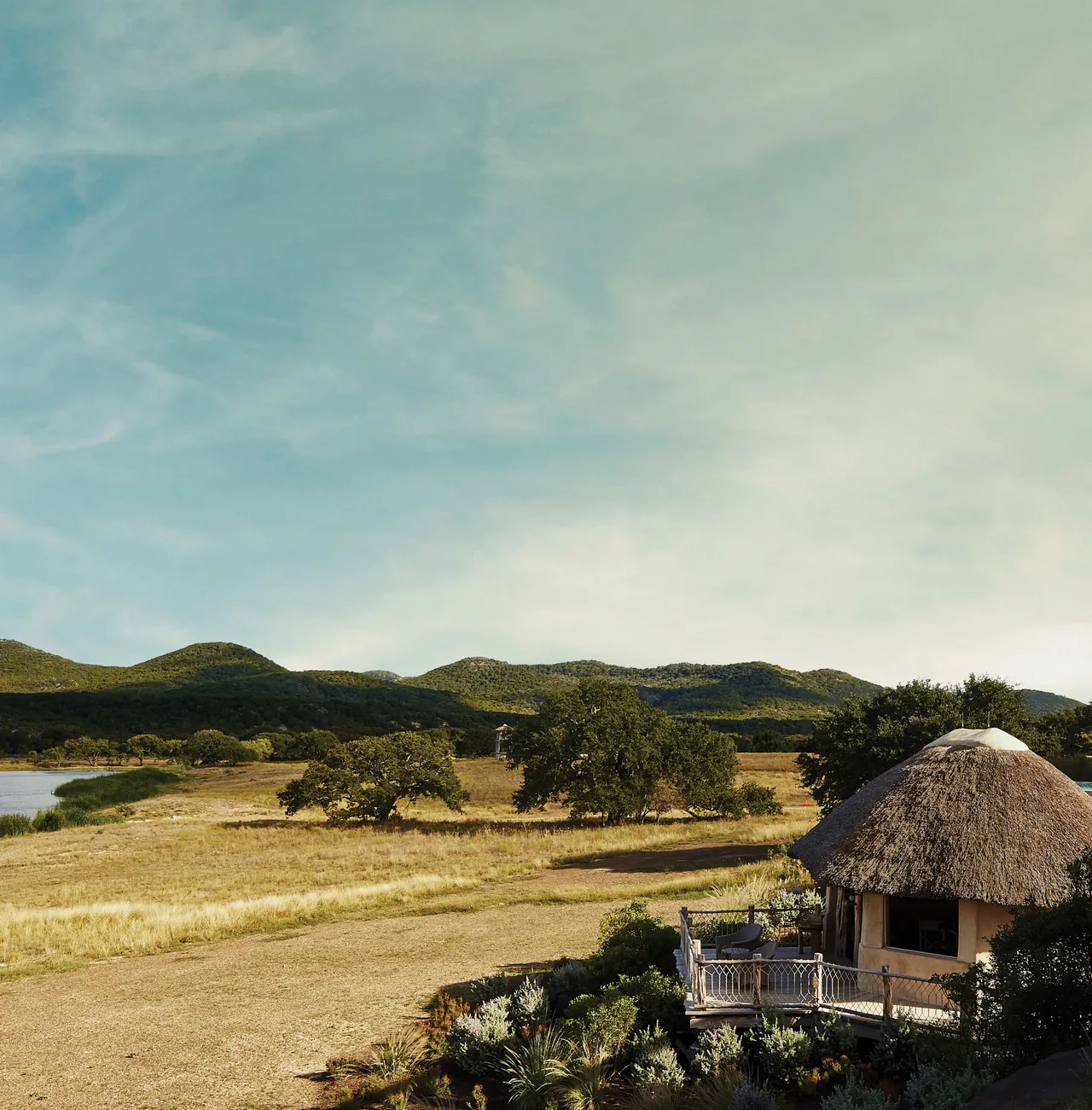 A round hut with a thatched roof and wooden railing sits at the edge of a dry field with hills in the background.