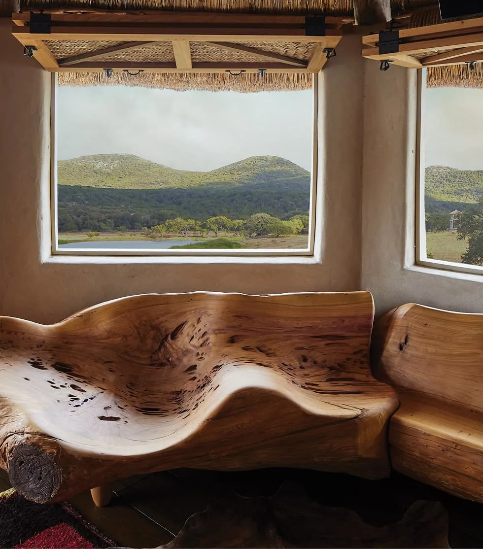 Sculpted wooden benches inside a room with plaster walls and large windows overlooking a hill landscape.