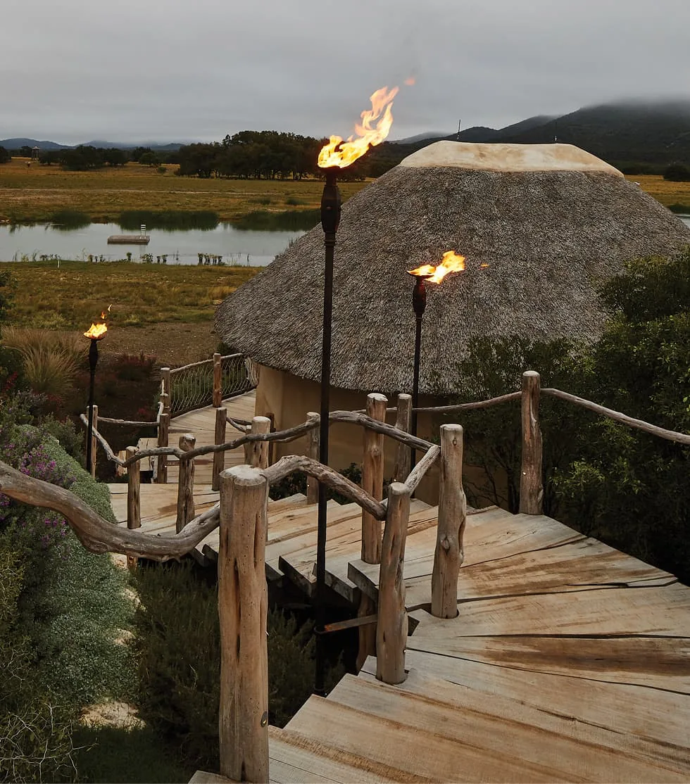 Wooden staircase with rustic branch railing and flaming tiki torches beside a thatched roof building near a pond.