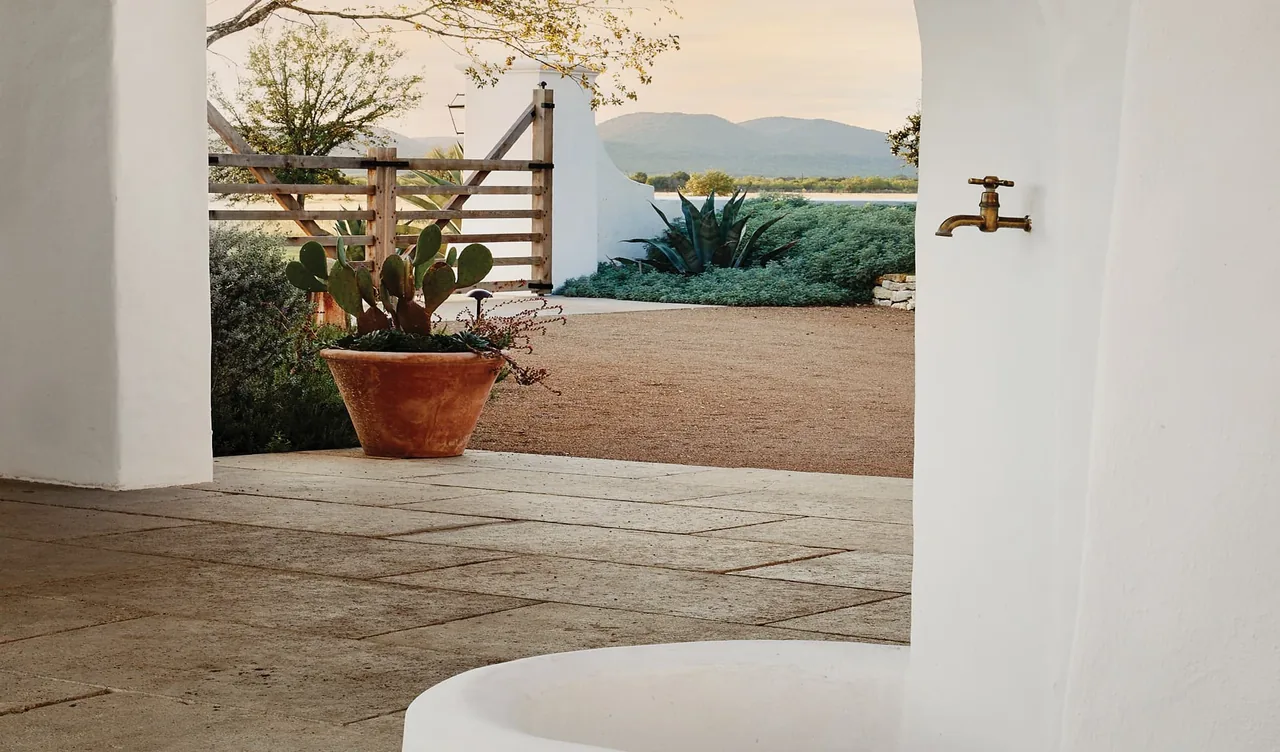 Outdoor view through a curved white plaster wall with brass faucet, stone tile floor, and desert landscaping at Rancho Sabino Grande.