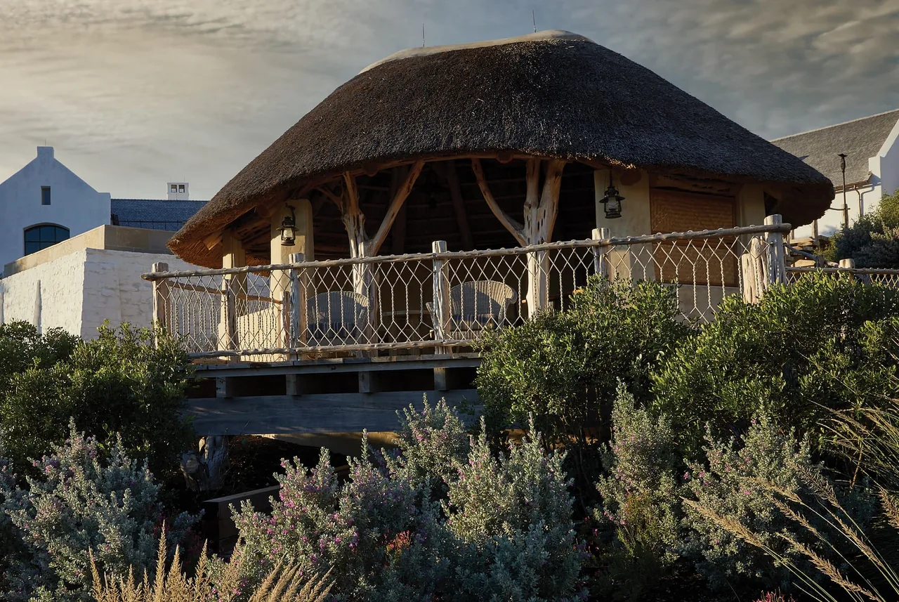 Thatched roof pavilion with natural wood supports and rope railing sits elevated amidst lush plants.