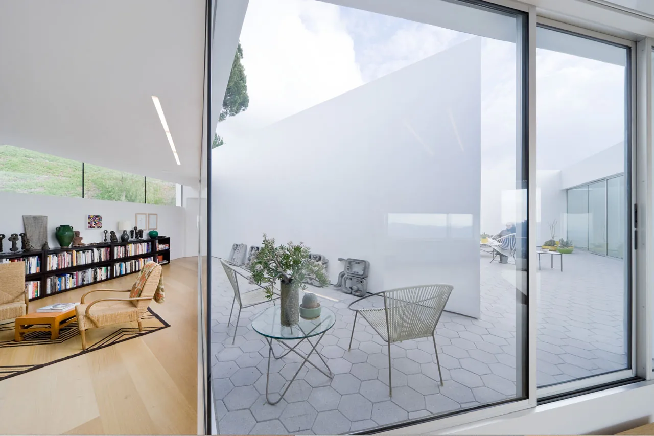 Living room with wooden flooring and woven chairs opens through a sliding glass door to an outdoor patio with hexagonal tiles and white walls.