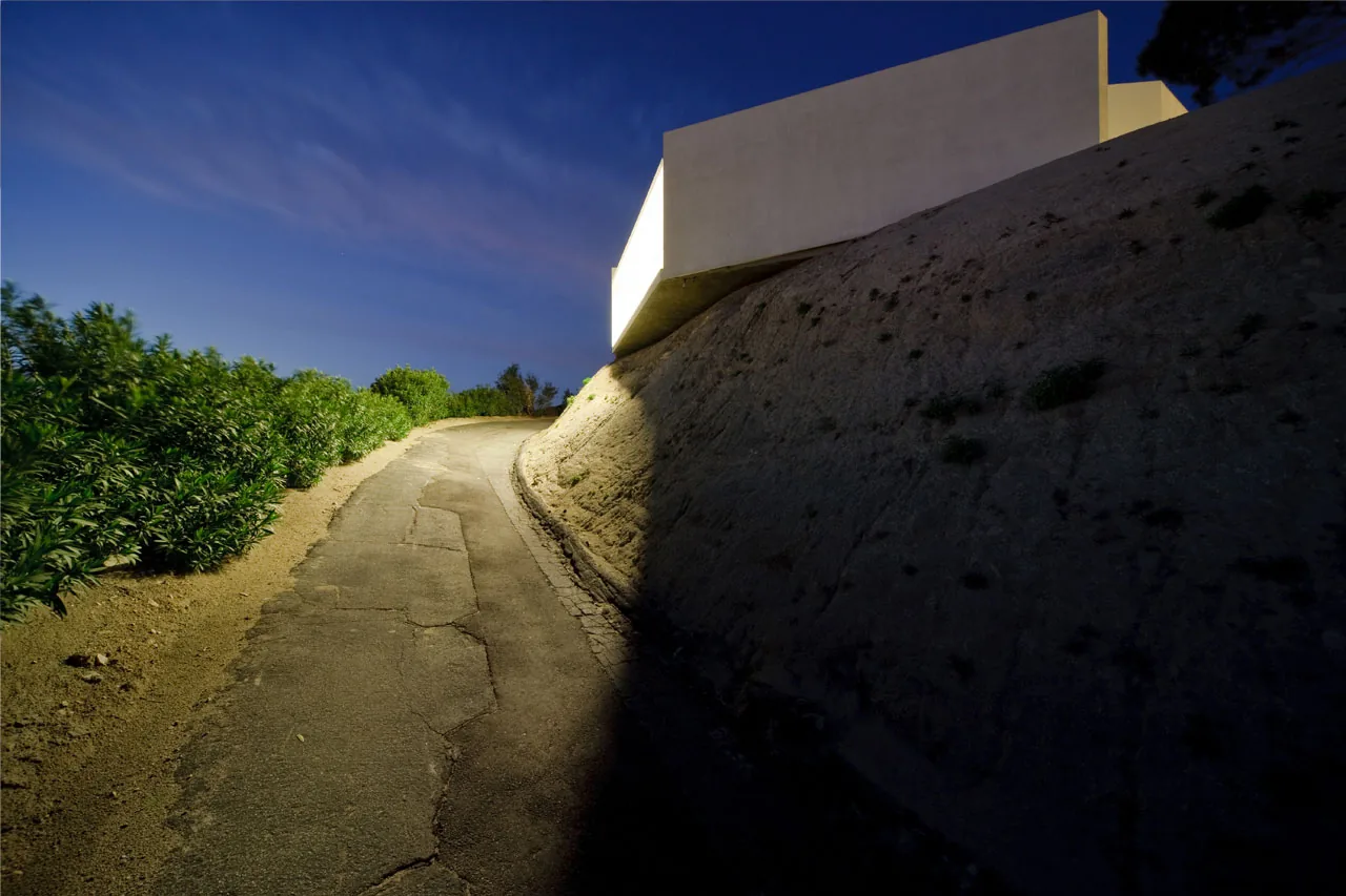 The Pittman Dowell residence features a minimalist white exterior built into a hillside, shown here at dusk with surrounding vegetation.