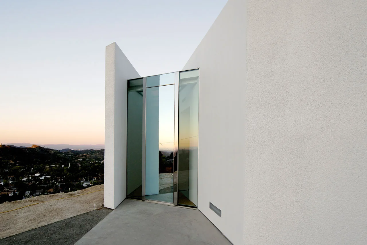 Corner entrance of the Pittman Dowell residence features a reflective glass pivot door and white stucco walls overlooking a landscape.