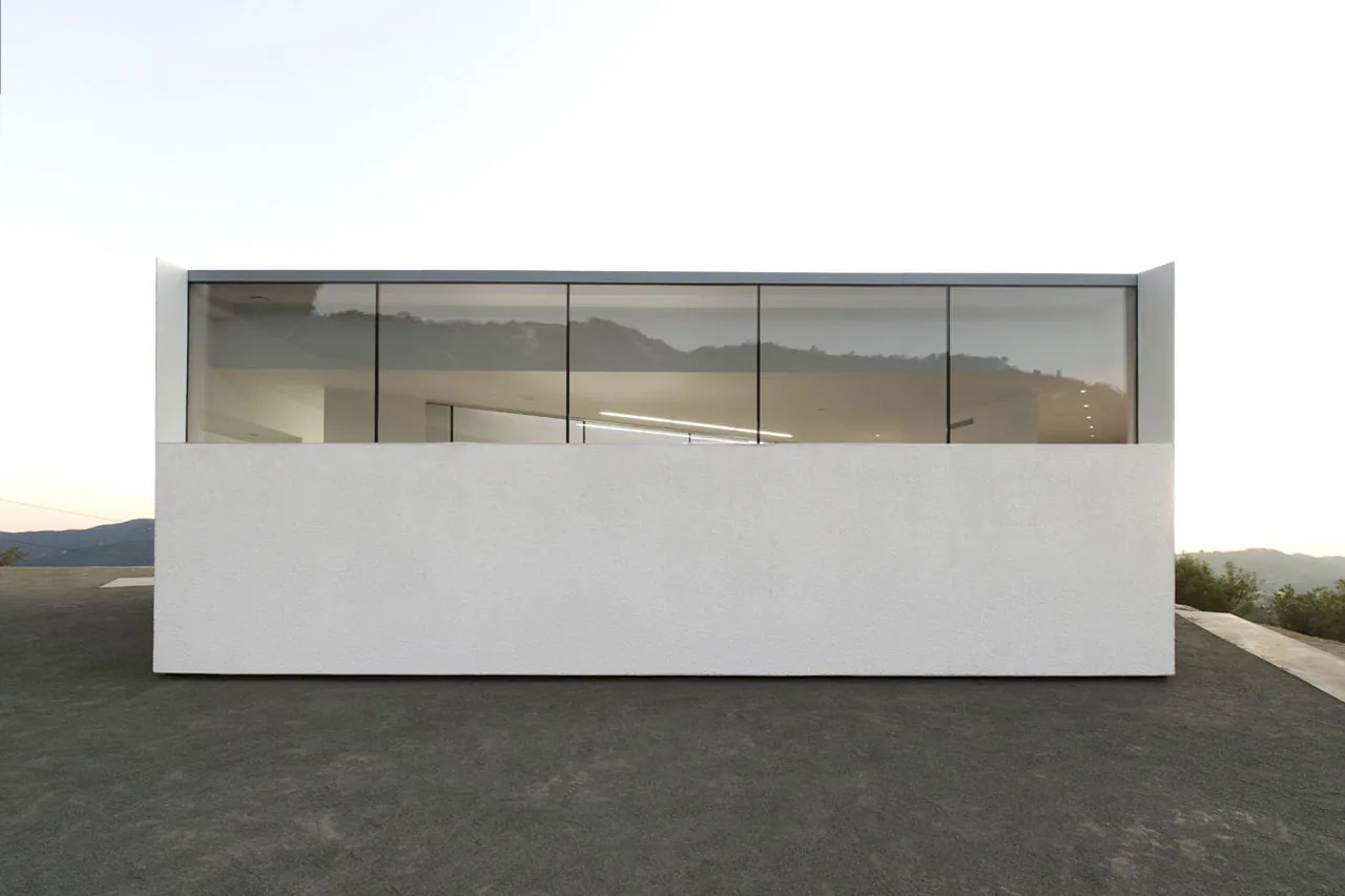 The Pittman Dowell residence features a minimalist white stucco facade with floor-to-ceiling reflective glass windows overlooking a hillside.