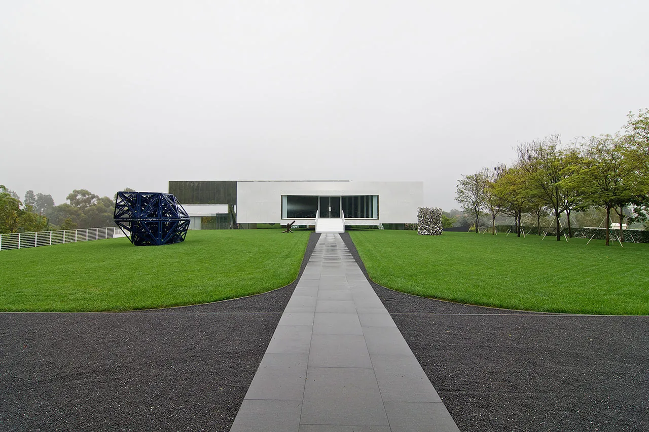 A minimalist white rectangular house with flat roof sits on a manicured lawn with modern sculptures in front.