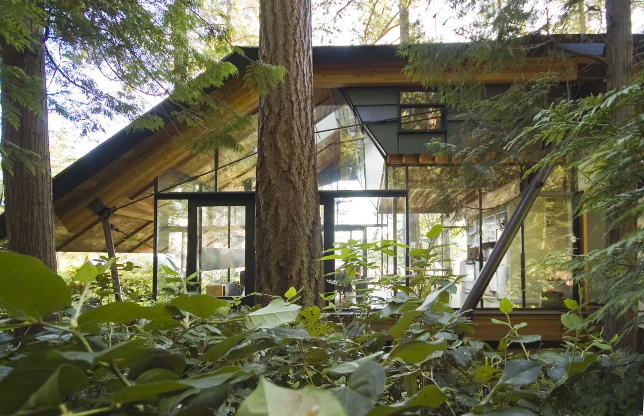 The glass cabin features floor-to-ceiling glazing with a gable roof, set in a wooded environment.