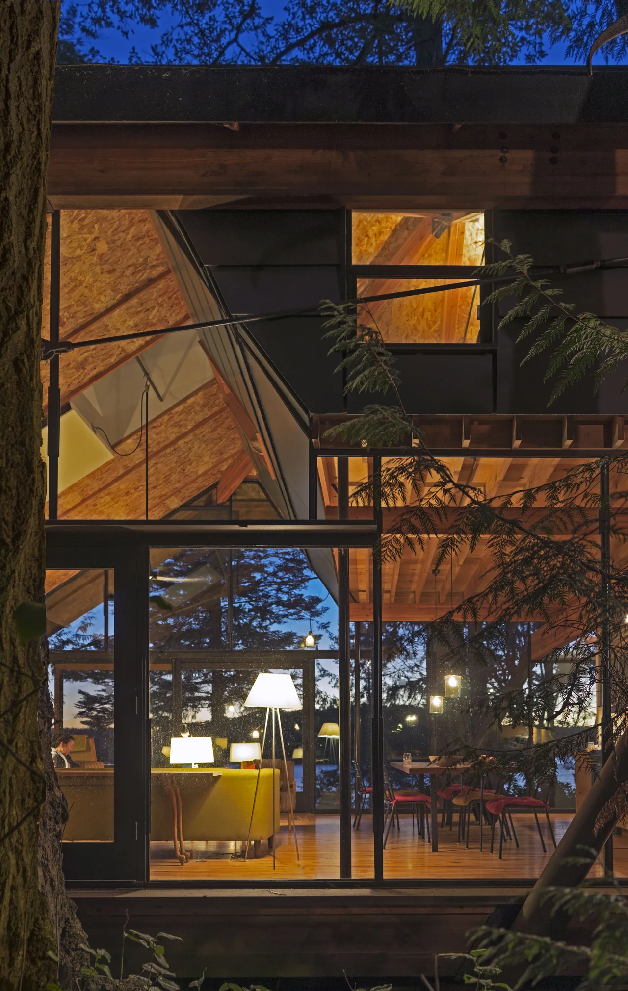 Glass cabin showing the illuminated interior with living and dining spaces, featuring wood floors and exposed ceiling beams