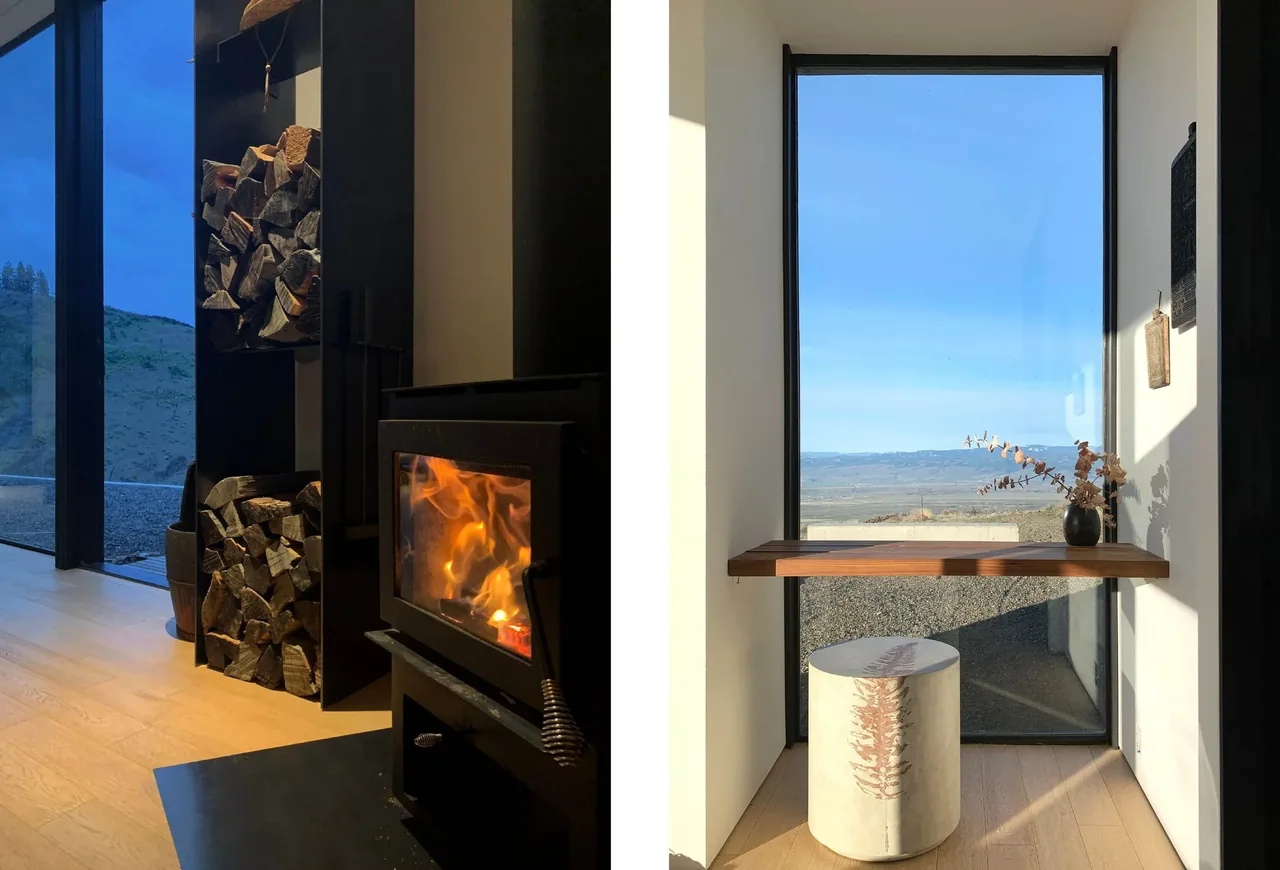 Modern interior with a wood stove next to floor-to-ceiling glazing and a small wood table by a large window overlooking the landscape.