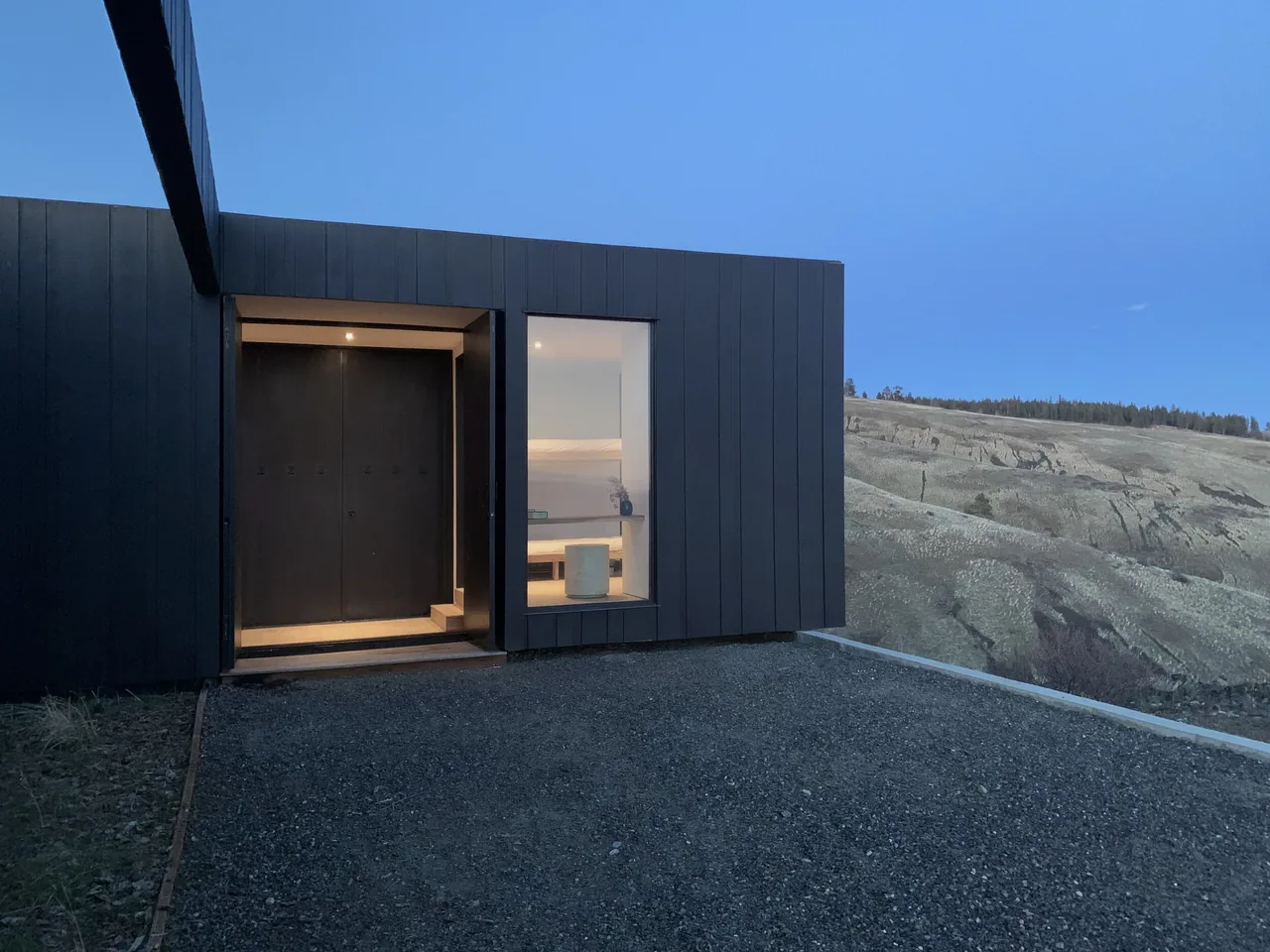 Basalt Canyon Shelter features black vertical siding with entry door and large window facing a hilltop landscape near sunset.