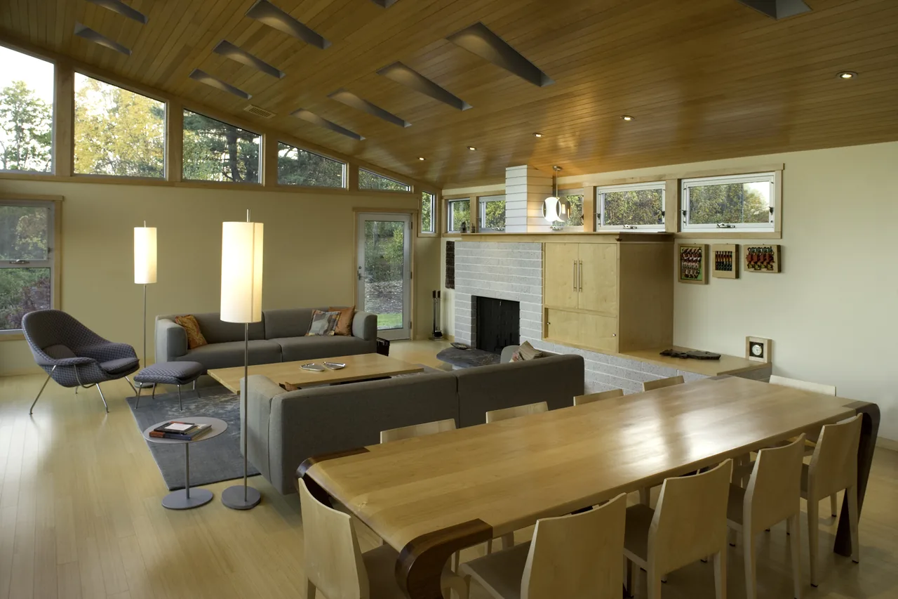 Open living and dining space with a wood-paneled sloped ceiling, floor lamps, and a fireplace built into a light brick wall.