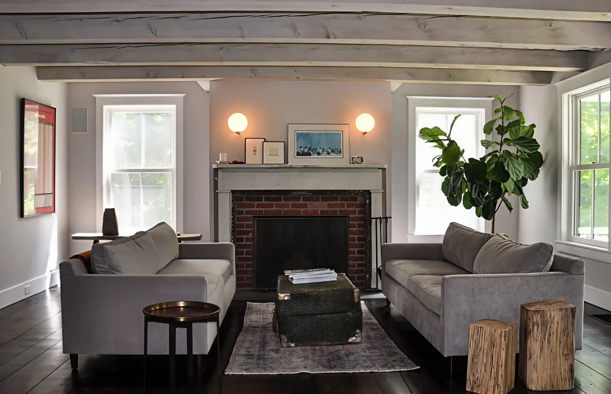 Living room of HUDSON VALLEY EXTREME MAKEOVER with brick fireplace, exposed beams, and symmetrical window placement.