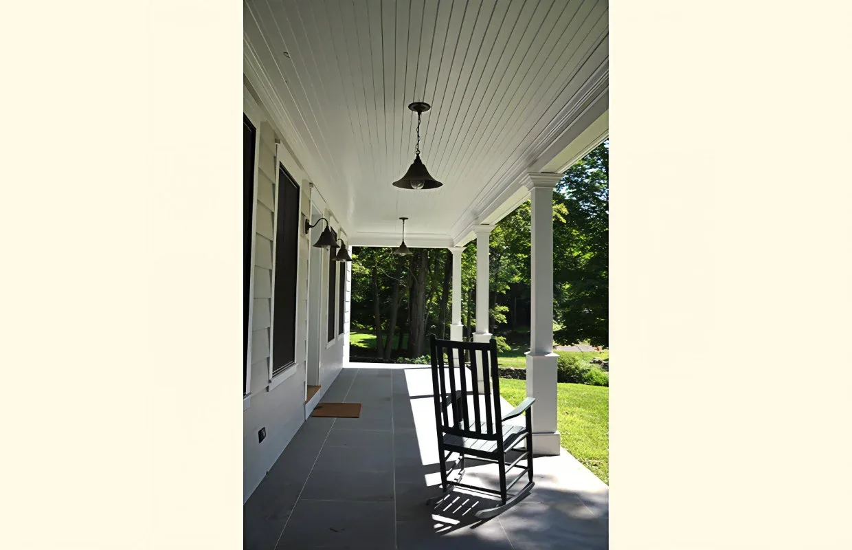 Covered front porch with white paneled ceiling, square columns, black rocking chair, and hanging pendant lights.