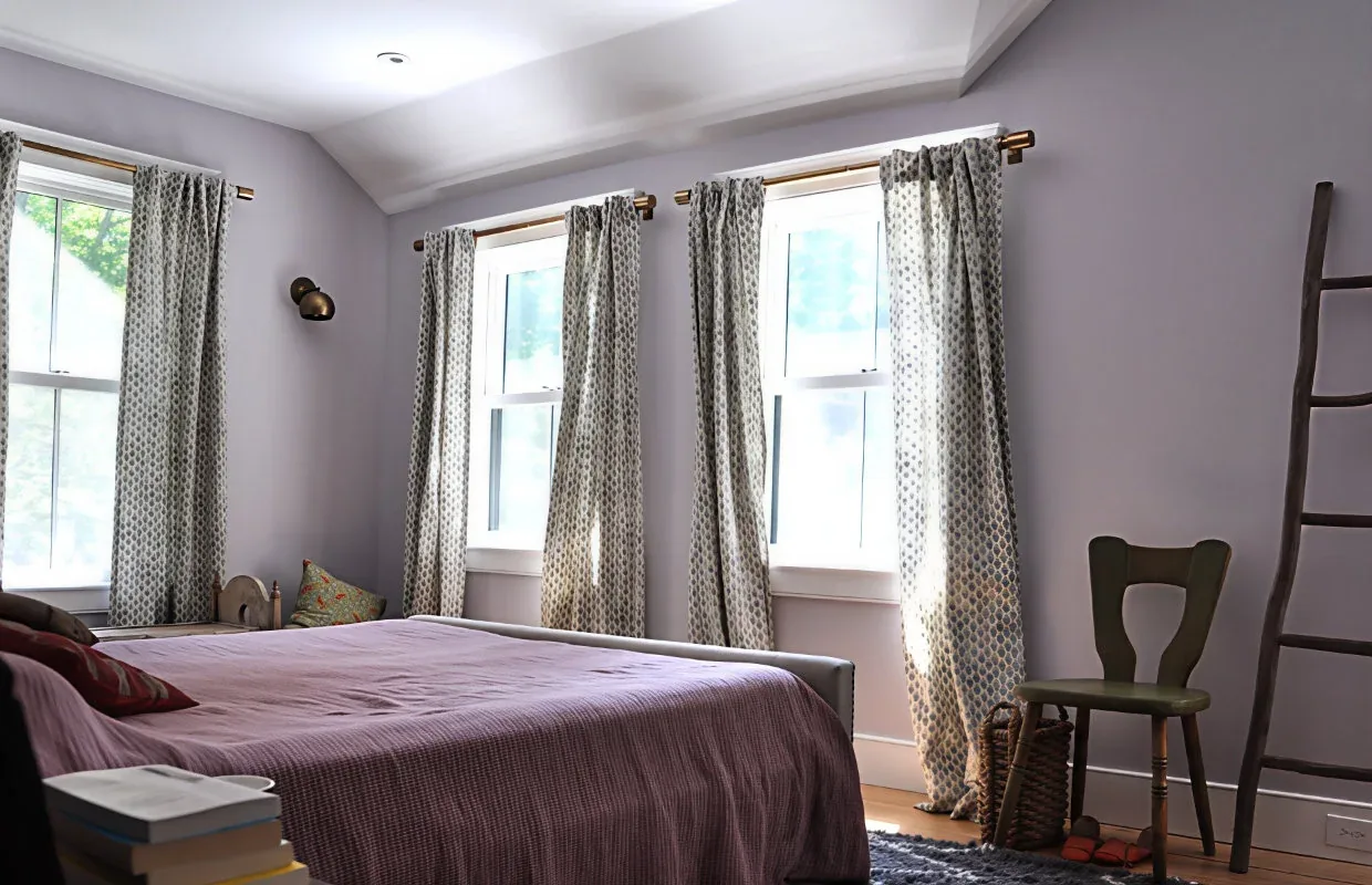 Bedroom featuring symmetrical double-hung windows with patterned curtains and a simple bed with mauve bedding.