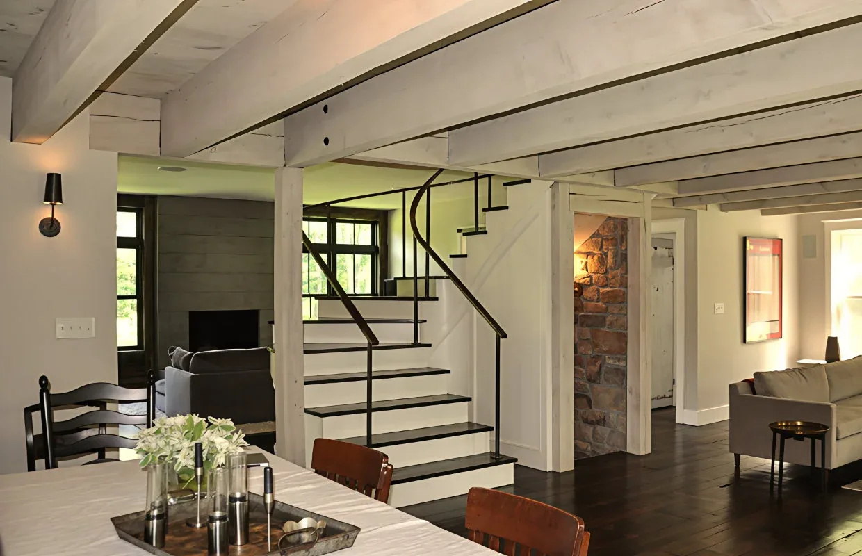 Open interior with exposed wood ceiling beams, black metal stair railing, and mixed dining and living areas with dark flooring.