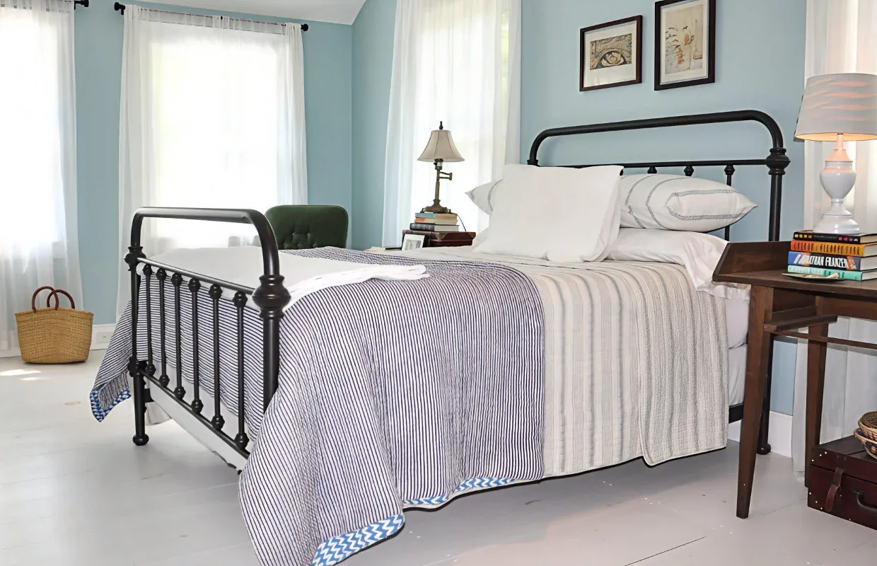 This bedroom features a black metal bed frame and pale blue walls with natural light through white curtains.