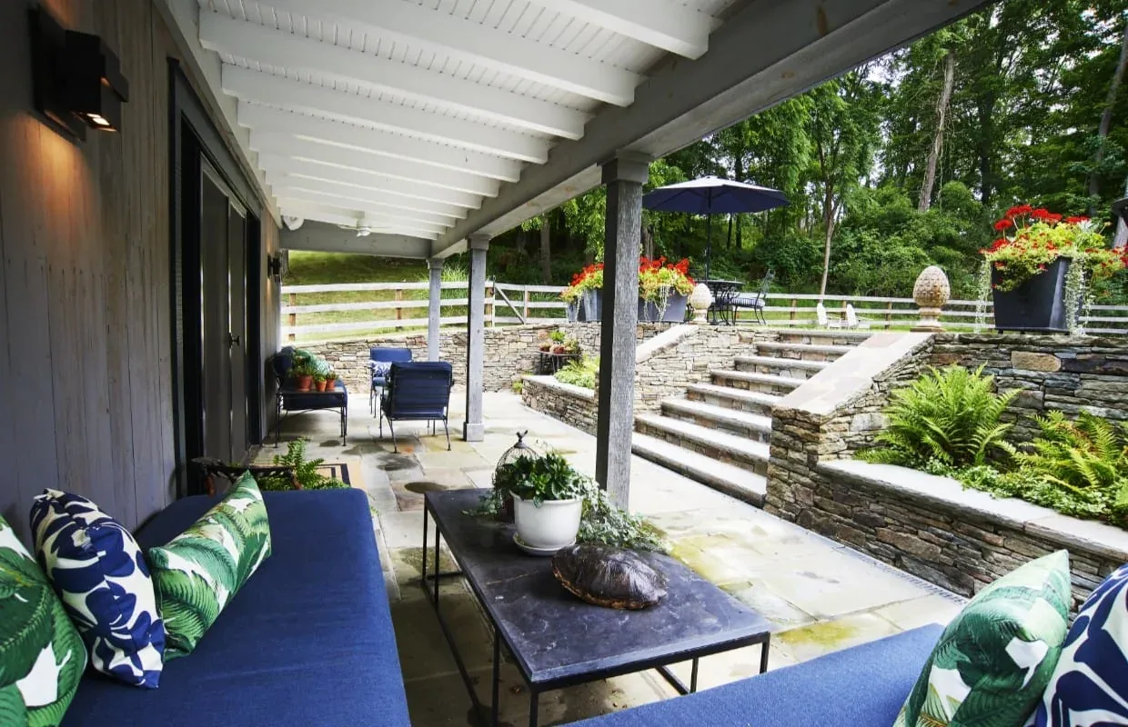 A covered patio at Frog Pond Farm features stone flooring, seating with blue cushions, and stone steps leading to an outdoor dining area with an umbrella.
