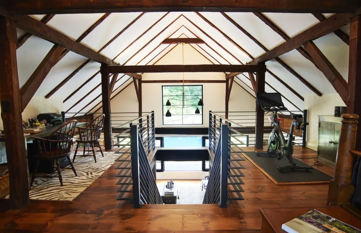 Loft with exposed wood beam ceiling, zebra-pattern rug, wood desk, and exercise bike overlooking the lower level through railing.