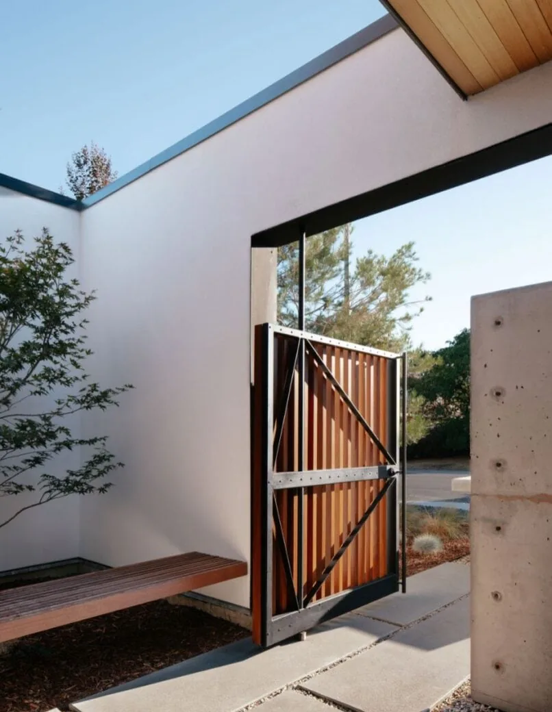 Courtyard gate with vertical wood slats and black metal framing beside a white stucco wall and wood bench.