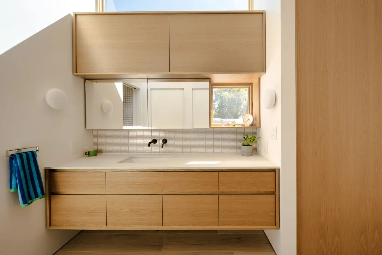 A bathroom vanity with light wood drawers and upper cabinets features a white countertop, wall-mounted faucet, and a small window with natural light.
