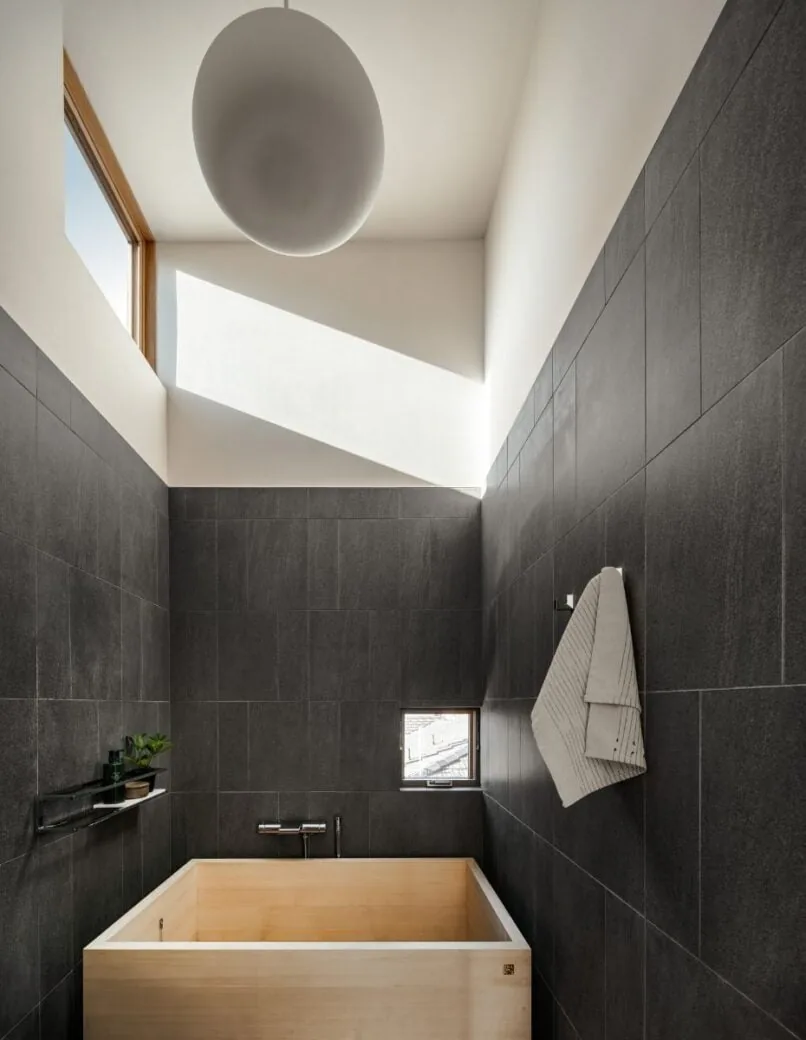 The bathroom features dark tiled walls, a light wood soaking tub, a small high window, and a towel hanging on the wall.